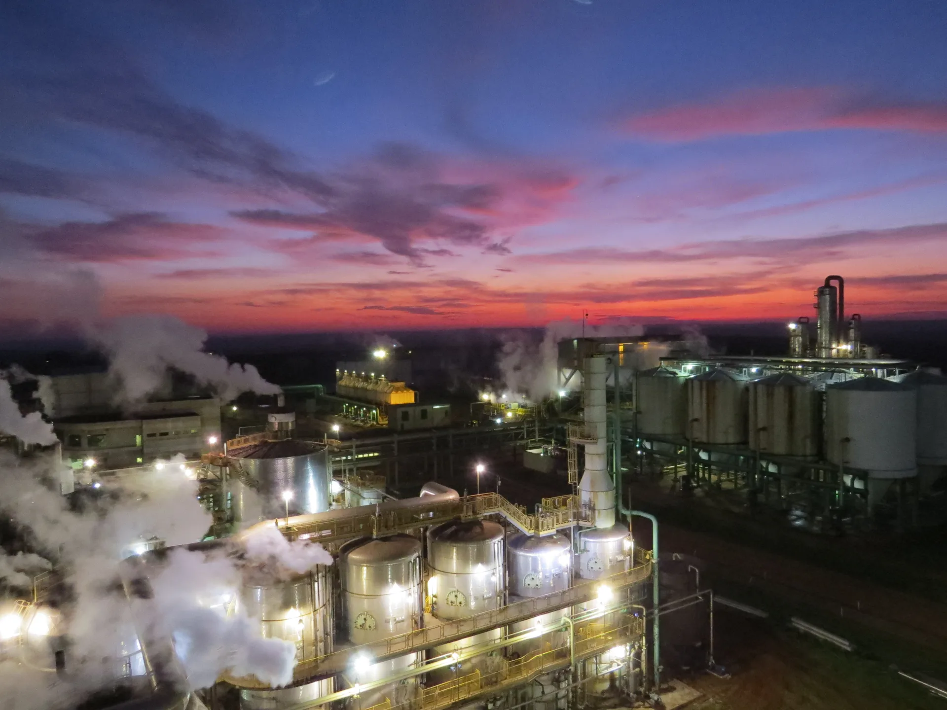 Vista noturna do complexo industrial da Usina Da Mata ao entardecer, com vapor das colunas de processo